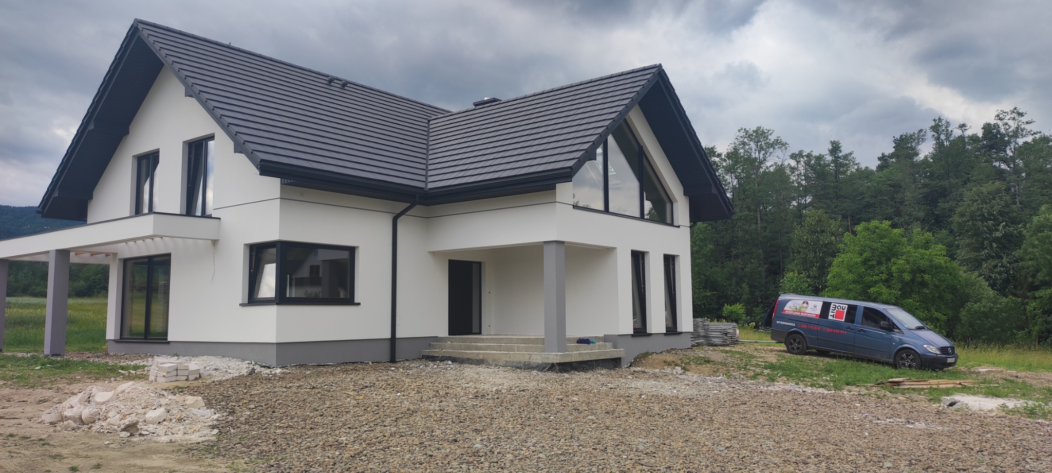 Nowoczesny, dwukondygnacyjny dom z białymi ścianami i ciemną dachówką, widoczny z zewnątrz, w trakcie prac wykończeniowych, z zaparkowanym samochodem dostawczym firmy budowlanej na posesji.