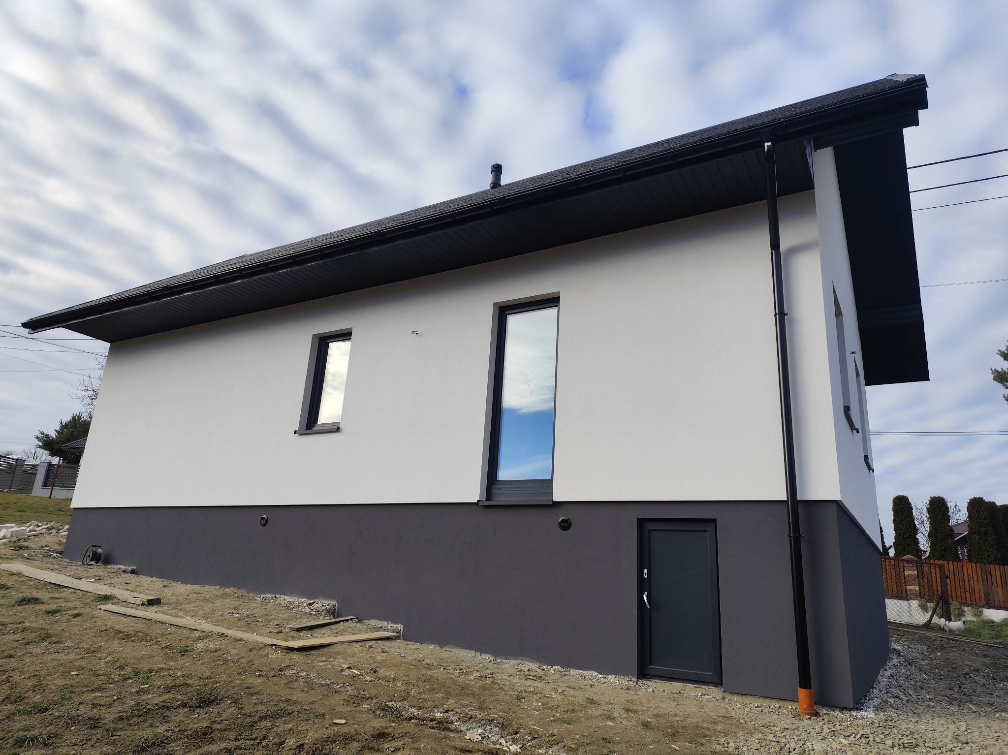 Dwukolorowy dom z oknami i drzwiami w ciemnej ramie, świeżo po tynkowaniu. Widoczne fragmenty terenu wokół budynku. Pochmurne niebo odbija się w szybach.