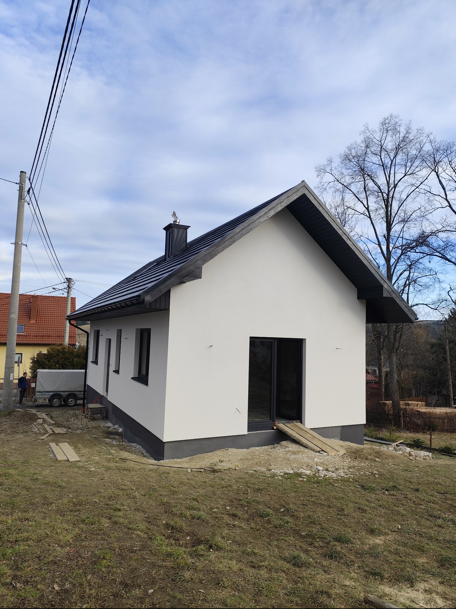 Biały dom z ciemną stolarką i dachem, widok z zewnątrz. Świeżo otynkowana elewacja, teren wokół domu jeszcze niezagospodarowany. Widoczne deski prowadzące do wejścia.
