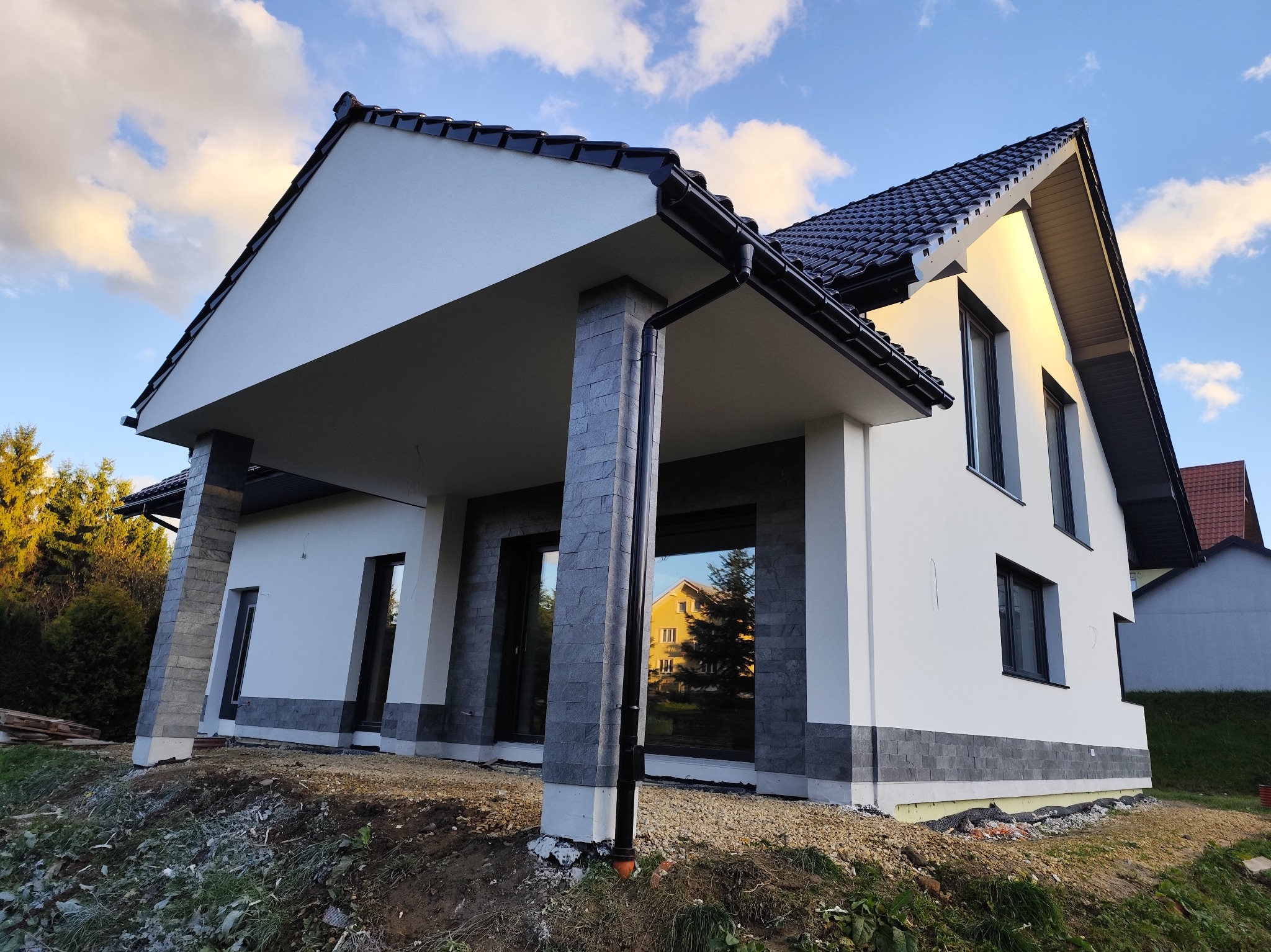 Nowoczesny dom z białymi ścianami i szarymi elementami dekoracyjnymi elewacji, widok z zewnątrz w słoneczny dzień. Widoczne okna i dach z czarną dachówką.