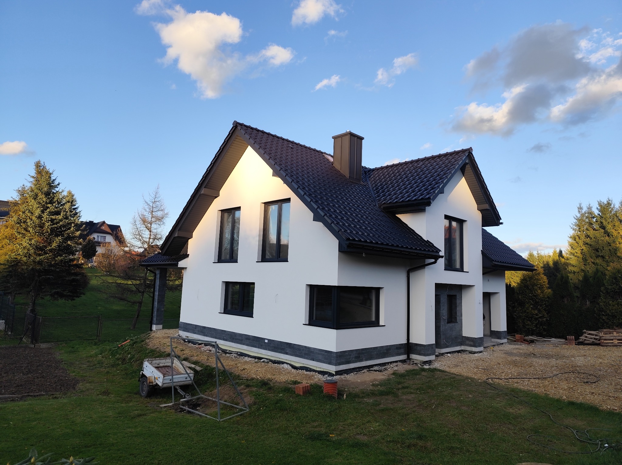 Nowoczesny dom jednorodzinny z białą elewacją i ciemną dachówką, widoczny z perspektywy ogrodu w słoneczny dzień. Uwagę zwraca wykończenie elewacji imitujące kamień.