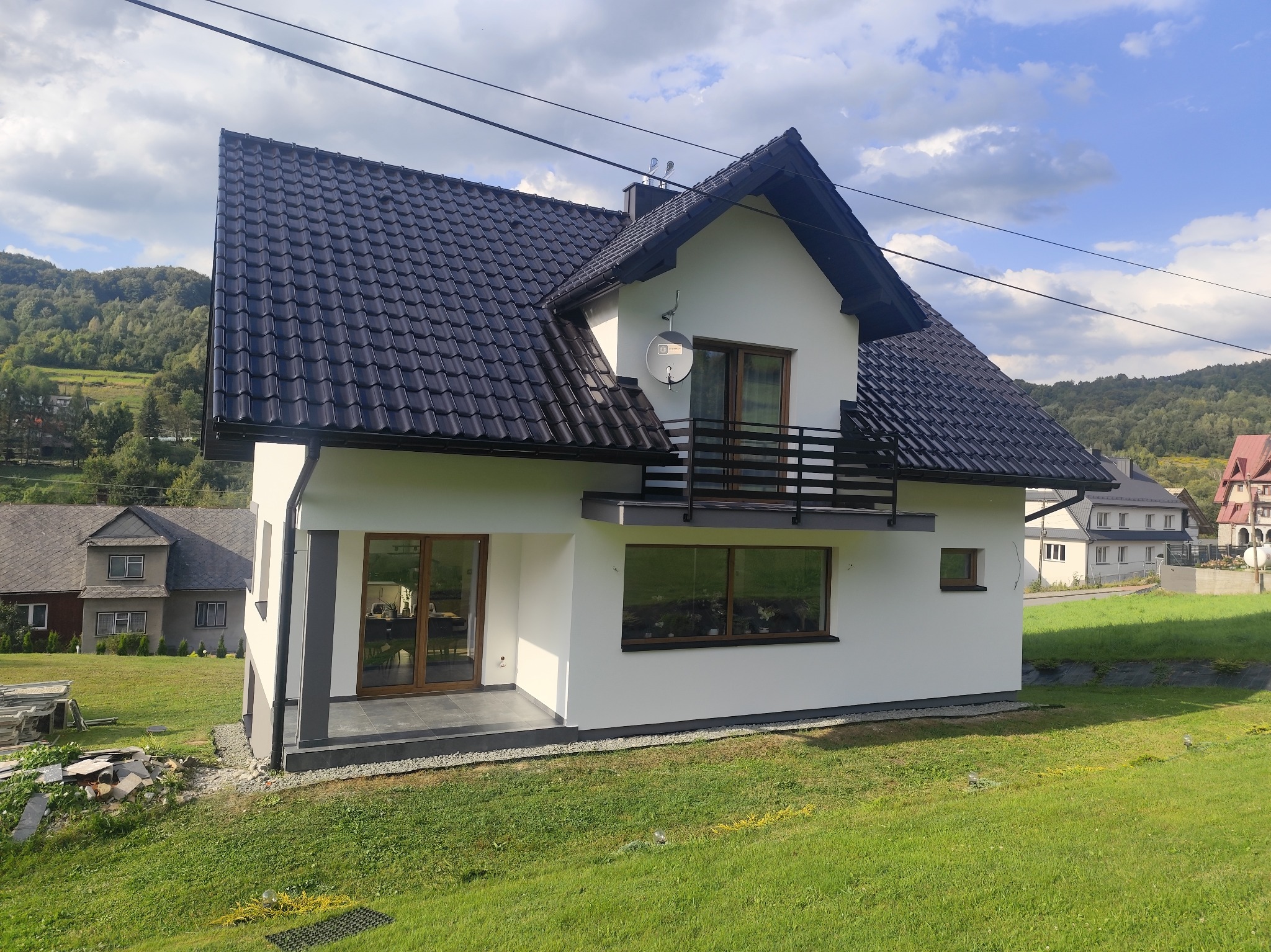 Biały, dwukondygnacyjny dom z ciemnym dachem na tle zielonego wzgórza. Widoczny balkon z czarną balustradą i satelita. Nowoczesna elewacja i okna z brązowymi ramami.