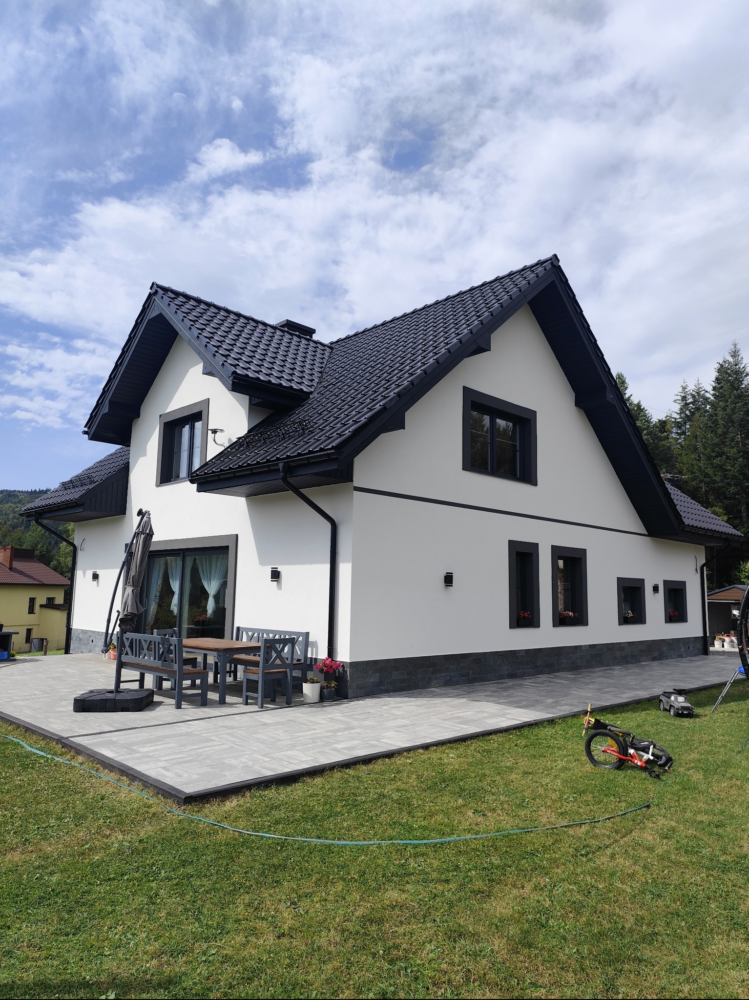 Biały dom z czarnym dachem i obramowaniami okien, otoczony zielonym trawnikiem i szarą kostką brukową. Widoczne meble ogrodowe i zabawki.