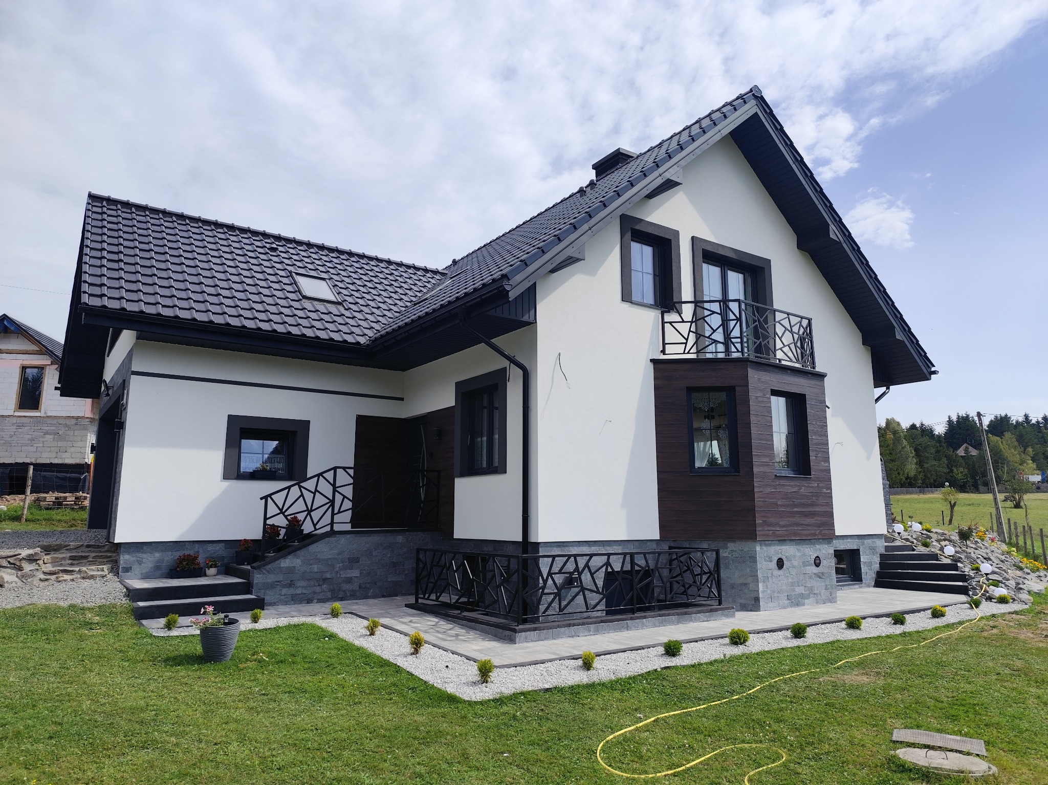 Nowoczesny dom z białą elewacją, czarną stolarką i dachem z dachówki. Widoczny balkon z geometryczną balustradą i kamienne wykończenie.
