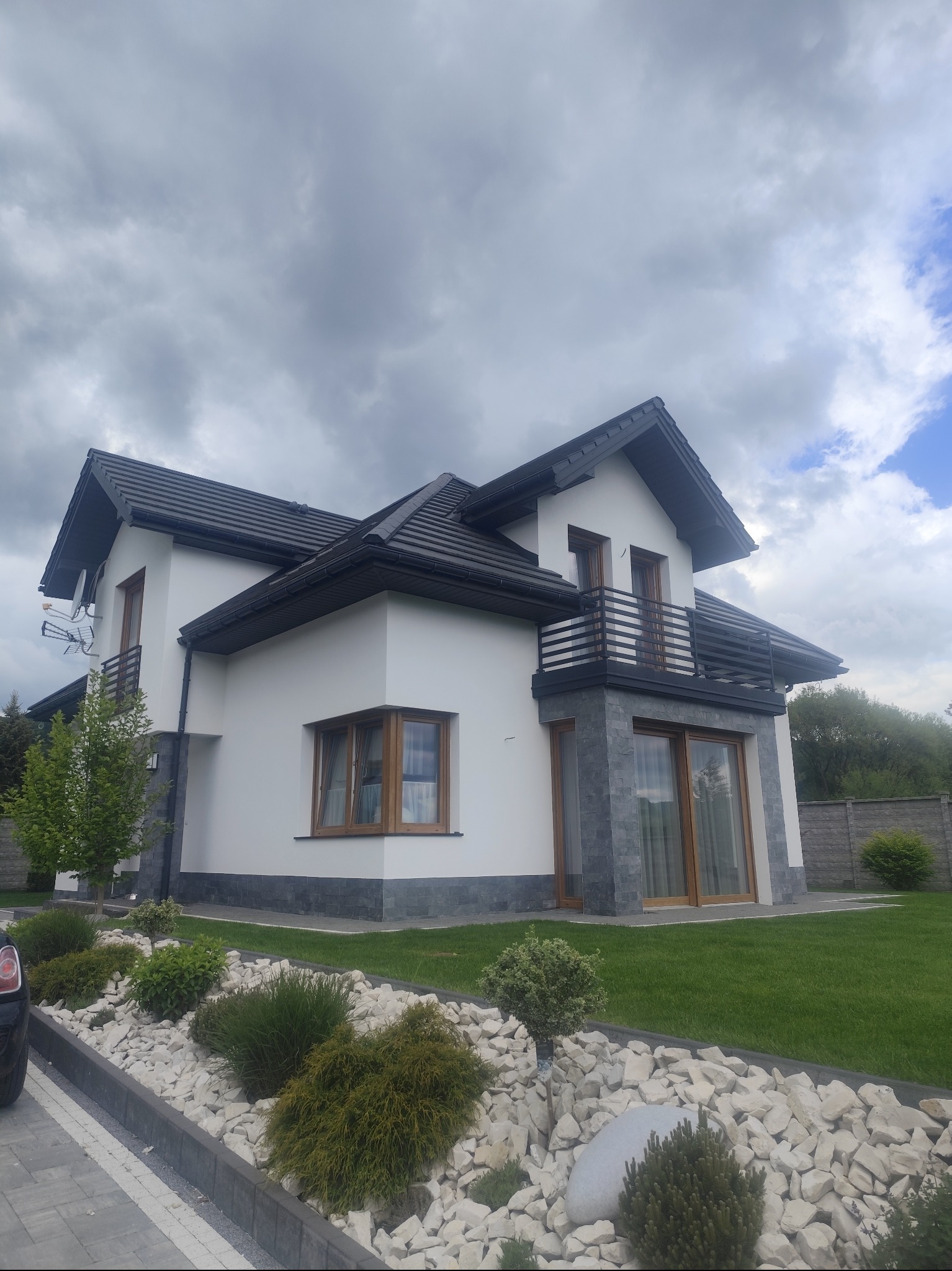 Nowoczesny dom jednorodzinny z białymi ścianami i ciemną dachówką, wykończony kamieniem elewacyjnym. Widok na zadbany ogród z ozdobnymi kamieniami i krzewami.