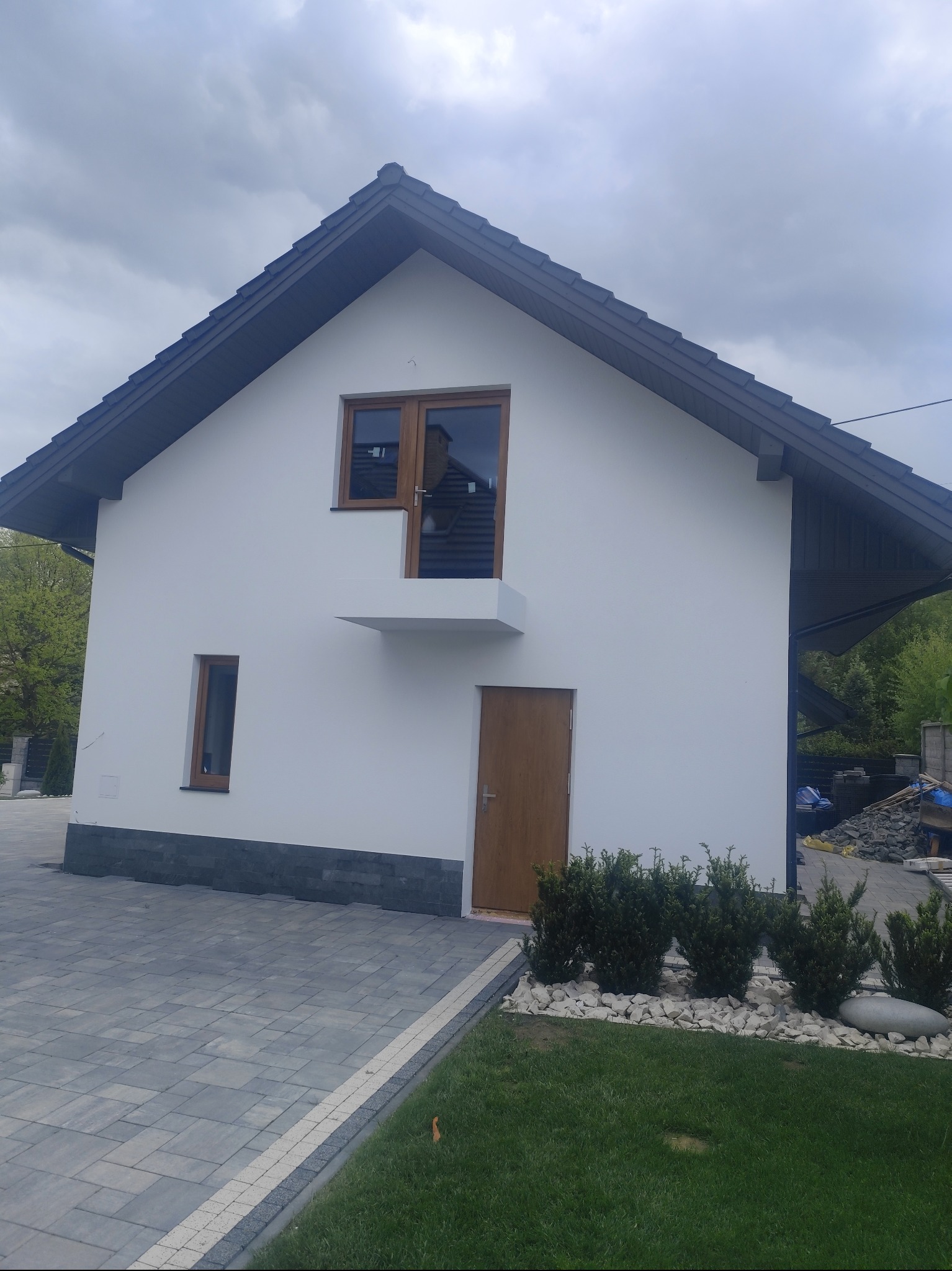 Biały dom jednorodzinny z ciemną dachówką i drewnianymi oknami, widok z poziomu chodnika. Elewacja po tynkowaniu, z elementami dekoracyjnymi z kamienia.