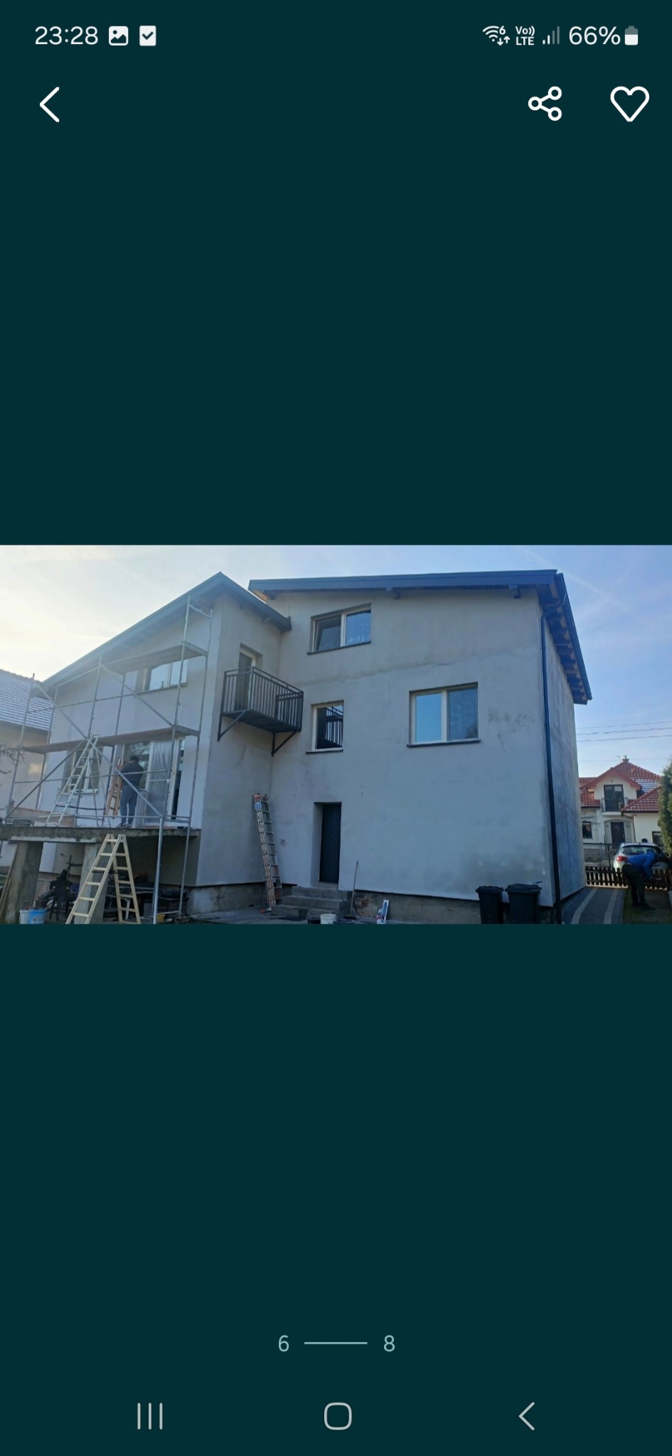 Dwupiętrowy dom w trakcie prac wykończeniowych elewacji, widoczne rusztowania, drabiny i pracownik w oknie, ściany częściowo pokryte szarą warstwą, balkon z czarną balustradą.