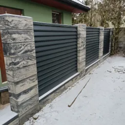 Nowoczesne ogrodzenie panelowe z grafitowymi przęsłami i słupkami z imitacji kamienia, częściowo zasypane śniegiem, na tle zielonego domu.