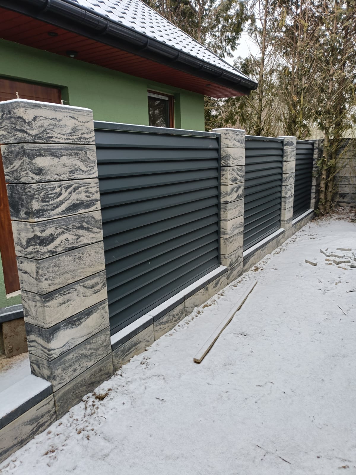 Nowoczesne ogrodzenie panelowe z grafitowymi przęsłami i słupkami z imitacji kamienia, częściowo zasypane śniegiem, na tle zielonego domu.