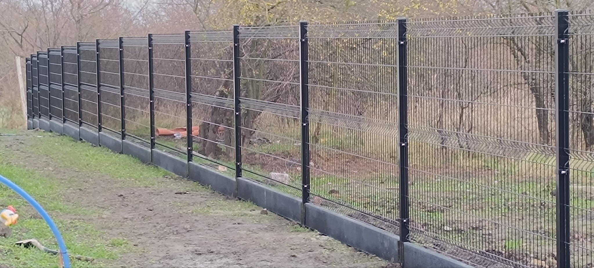 Długi, panelowy płot z podmurówką na trawiastym terenie, widoczny z boku. Część paneli z przesłonami. Tło stanowią drzewa bez liści i rury pcv.