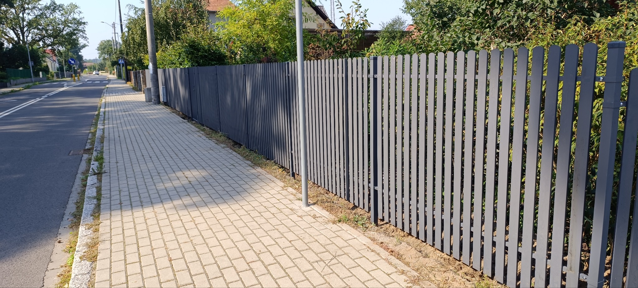 Długi, szary, panelowy płot z pionowych desek, oddzielający chodnik od posesji z zielenią. Widok na ulicę i domy w tle, w słoneczny dzień.