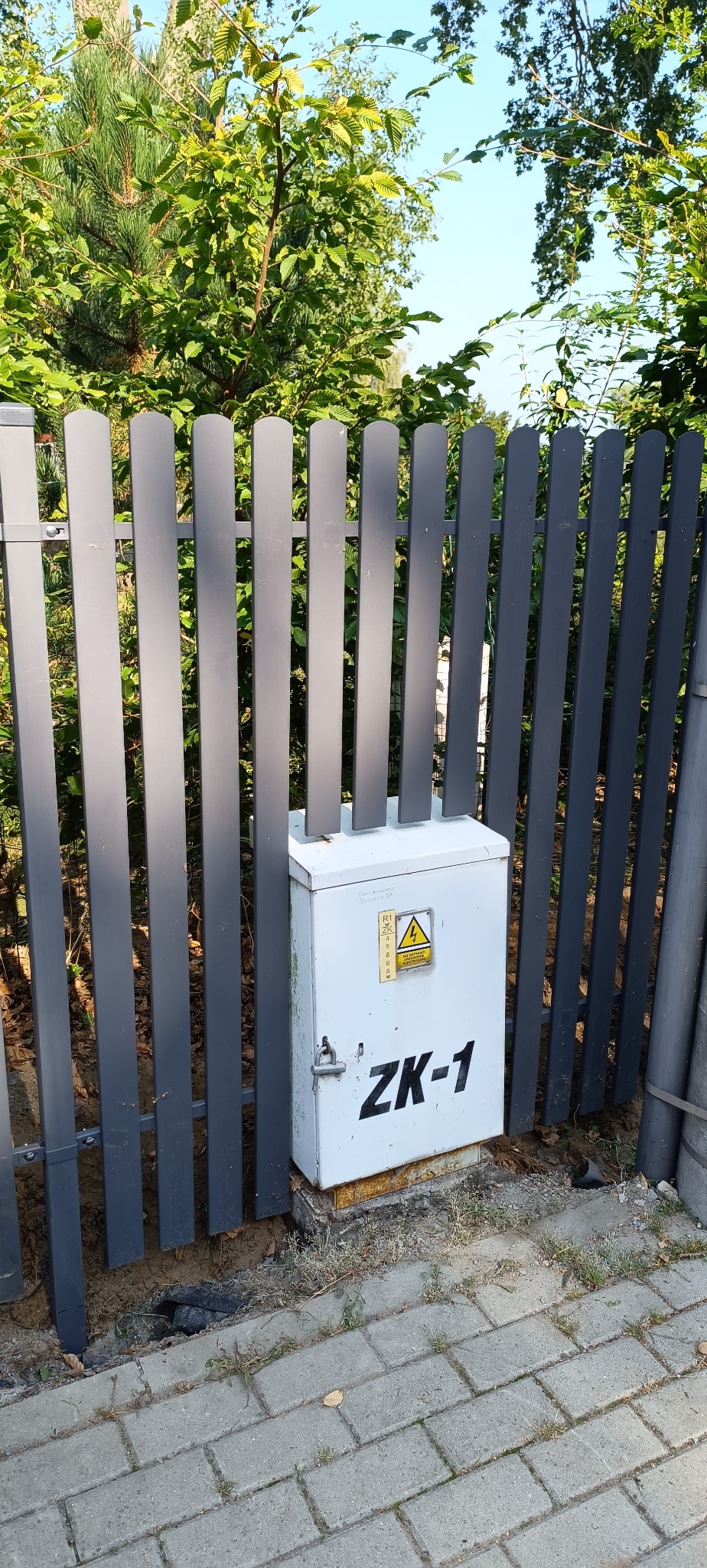 Szary, metalowy płot panelowy z białą skrzynką ZK-1 na tle zieleni. Widok z poziomu chodnika z kostki brukowej, w słoneczny dzień.