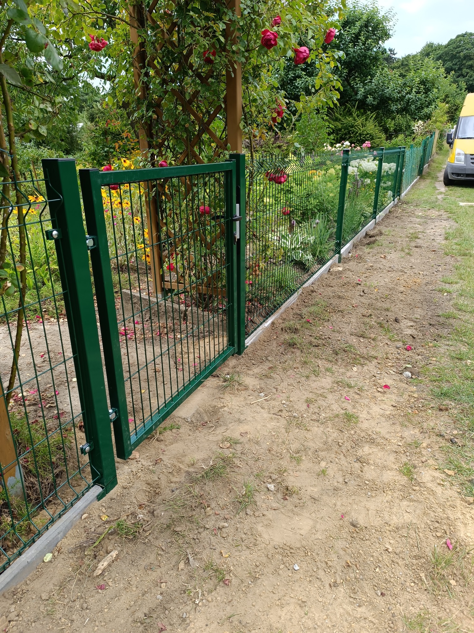 Nowoczesne ogrodzenie panelowe z zieloną bramką, otoczone kwitnącymi różami i bujną roślinnością. Żółty samochód dostawczy w tle.