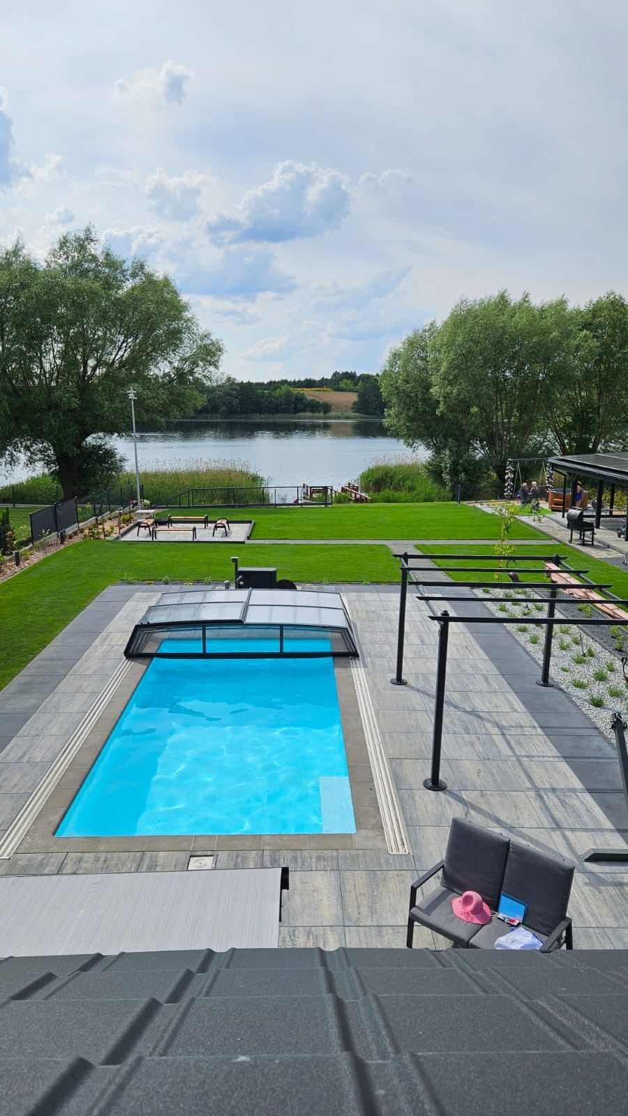 Prostokątny, błękitny basen z częściowo zasuniętą szarą roletą, otoczony szarą kostką brukową, z widokiem na jezioro i zielony trawnik; na pierwszym planie widoczny fragment dachu.