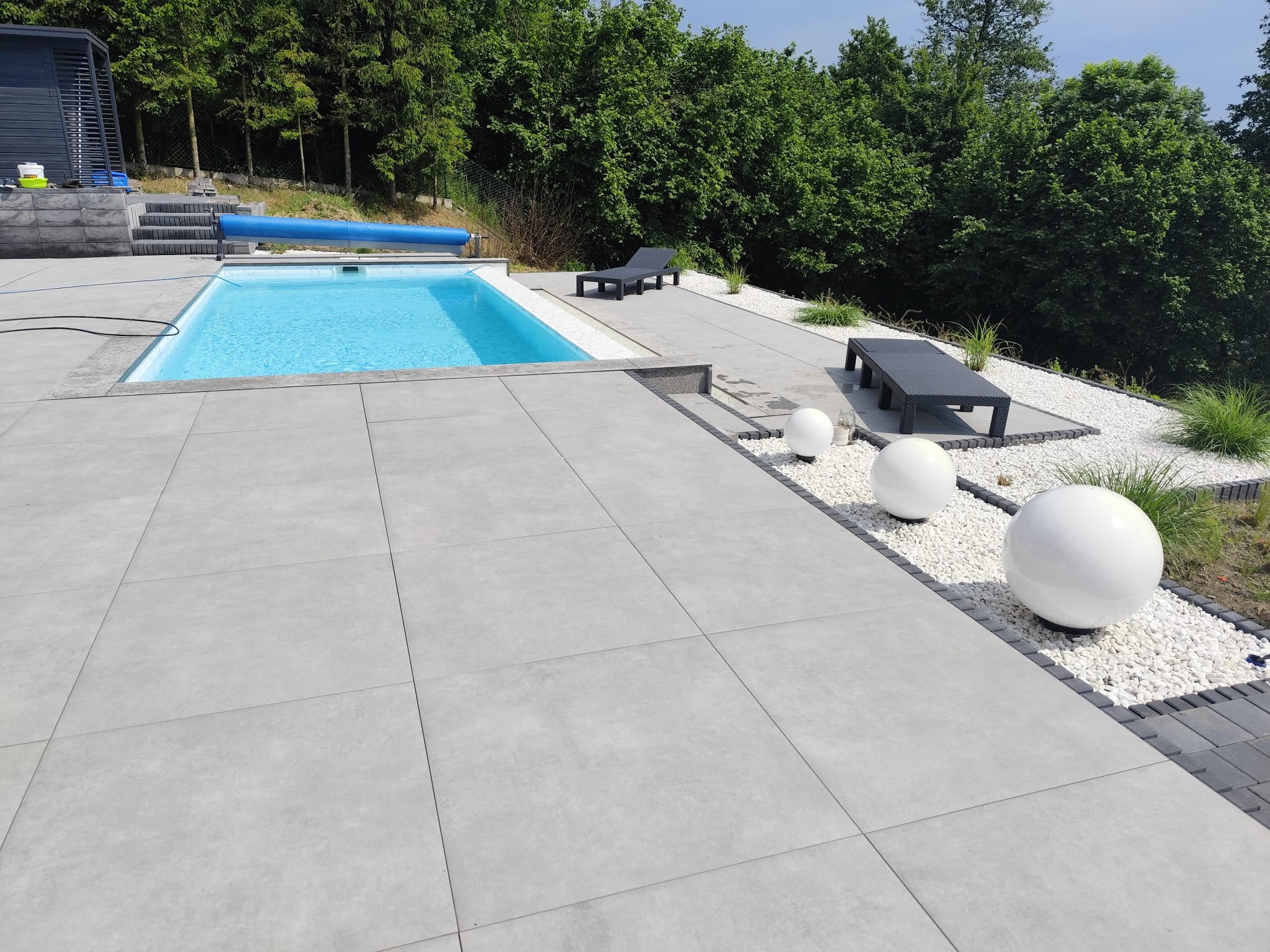 Nowoczesny basen ogrodowy z jasnymi płytami tarasowymi, białymi kulami dekoracyjnymi i leżakami na tle zieleni.