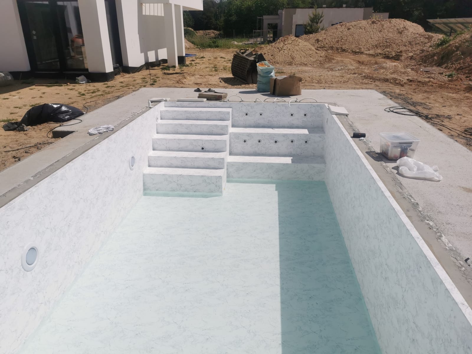 Wnętrze nowo wybudowanego basenu wyłożonego folią imitującą marmur, z wbudowanymi schodami i dyszami do hydromasażu, widoczne narzędzia i materiały budowlane na obrzeżach.