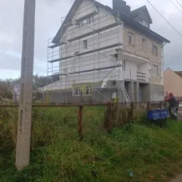 Dwupiętrowy dom w trakcie prac elewacyjnych: część ściany pokryta białym styropianem, reszta w surowym stanie. Rozstawione rusztowania, widoczne okna zabezpieczone folią. Z przodu zarośnięte...