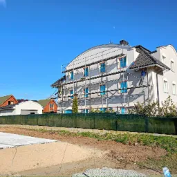 Dwupiętrowy dom z szarą elewacją w trakcie prac wykończeniowych, rusztowania zasłaniają część fasady, okna zabezpieczone folią, dwóch pracowników na rusztowaniach, błękitne niebo w tle.