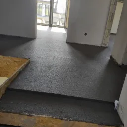 Świeżo wylana, ciemnoszara masa na podłodze w surowym wnętrzu, widoczne drewniane obramowanie schodka i niezabezpieczone otwory drzwiowe.