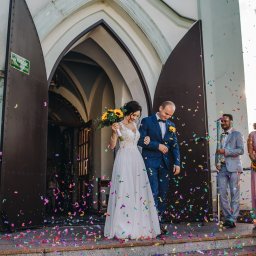 Natalia Jóźwiak - Para młoda opuszcza kościół w strugach kolorowego konfetti, panna młoda trzyma bukiet słoneczników, goście weselni obserwują i klaszczą.