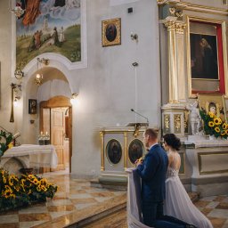 Natalia Jóźwiak - Para młoda klęczy przed ołtarzem w kościele, dekoracje z żółtych słoneczników, malowidła i ikony w tle.