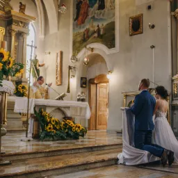 Para młoda klęczy przed ołtarzem w kościele, dekoracje z żółtych słoneczników, ksiądz w złotej szacie, marmurowa posadzka.