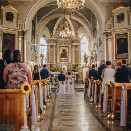 Natalia Jóźwiak - Para młoda klęczy przed ołtarzem w kościele podczas ceremonii ślubnej, widok zza pleców gości, ławki udekorowane słonecznikami i białymi wstążkami, świece wzdłuż przejścia.