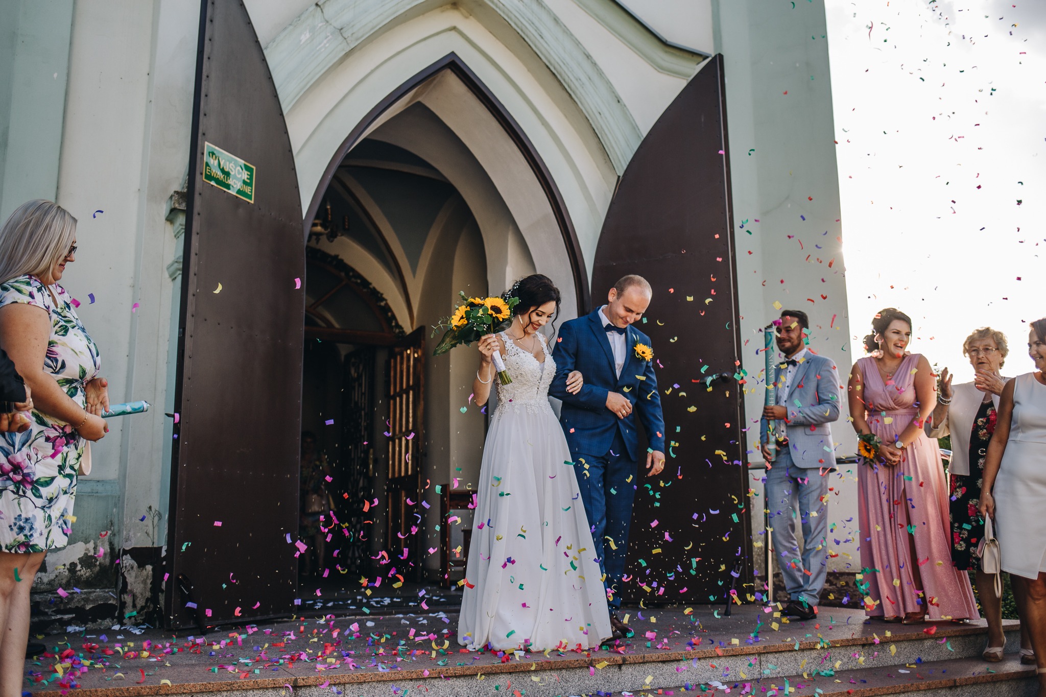 Para młoda opuszcza kościół w strugach kolorowego konfetti, panna młoda trzyma bukiet słoneczników, goście weselni obserwują i klaszczą.