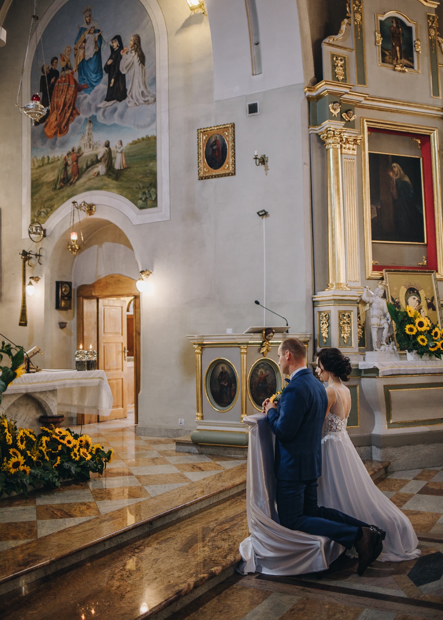 Para młoda klęczy przed ołtarzem w kościele, dekoracje z żółtych słoneczników, malowidła i ikony w tle.