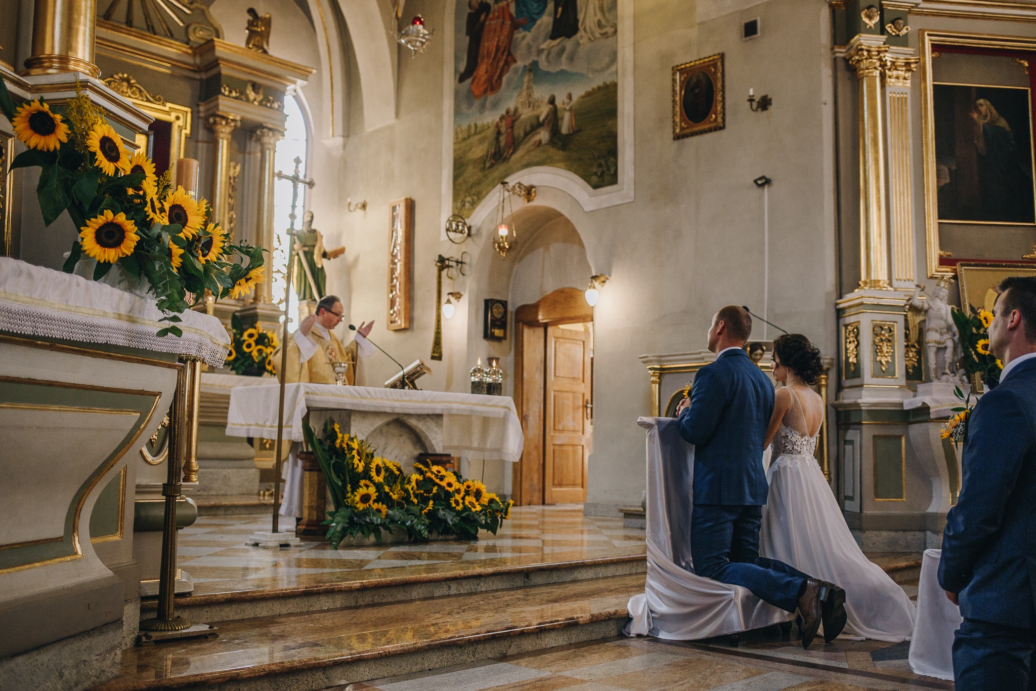 Para młoda klęczy przed ołtarzem w kościele, dekoracje z żółtych słoneczników, ksiądz w złotej szacie, marmurowa posadzka.