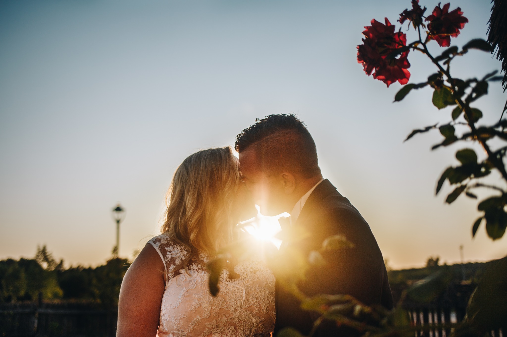 Para młoda całuje się w złotym świetle zachodzącego słońca, widoczne czerwone róże i latarnia w tle.