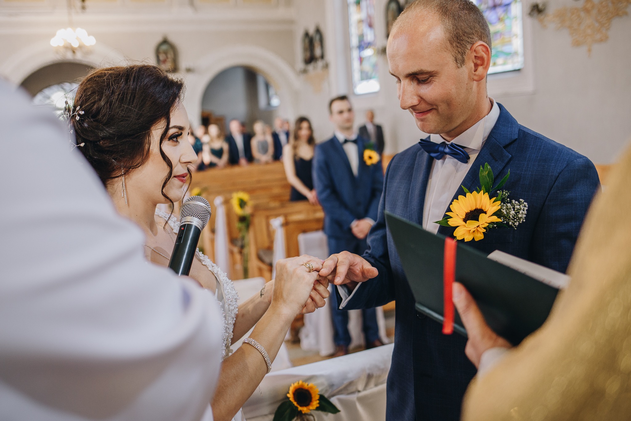 Wymiana obrączek podczas ceremonii ślubnej w kościele, panna młoda zakłada obrączkę panu młodemu, w tle goście i dekoracje z słoneczników.