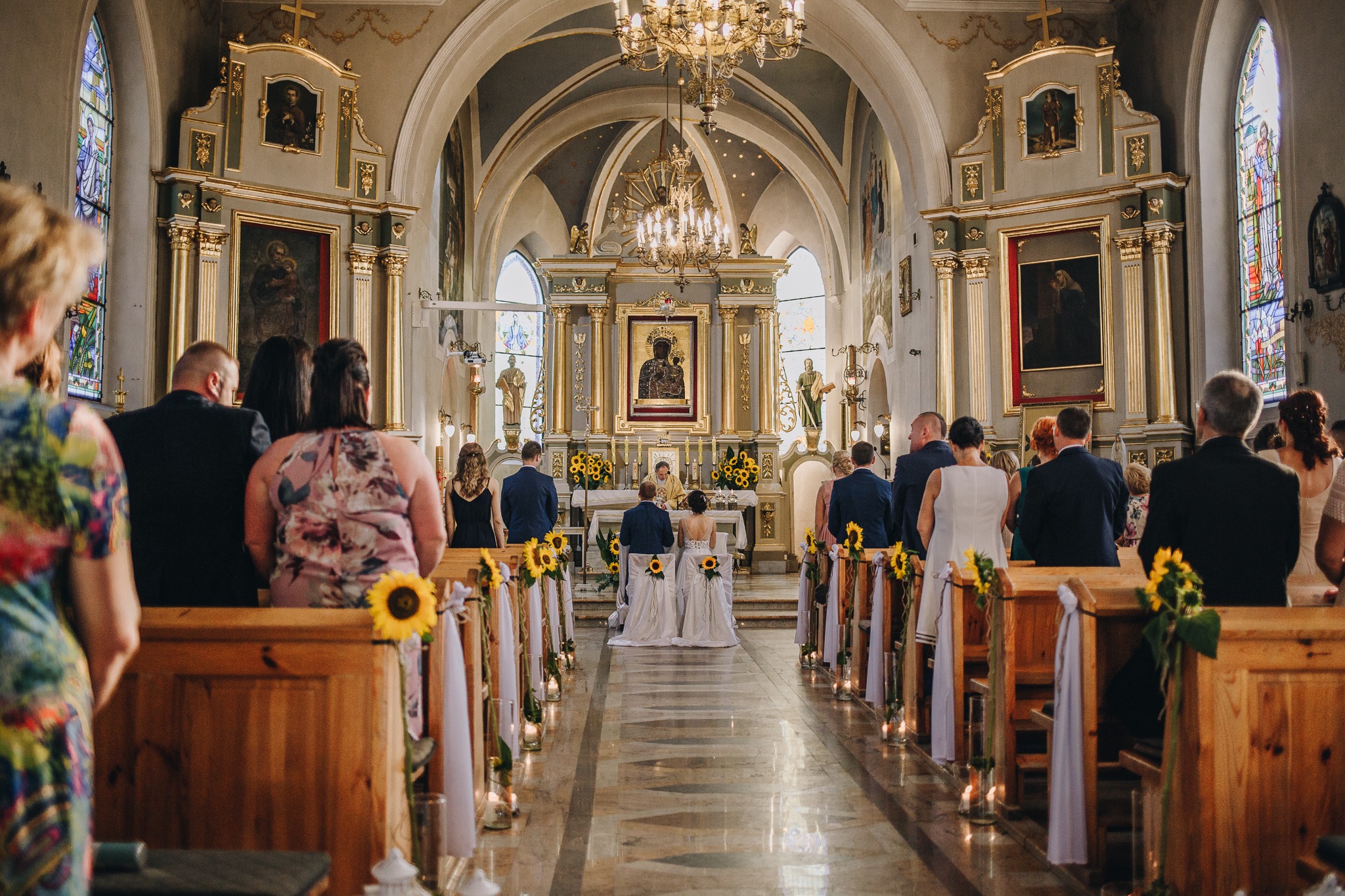 Para młoda klęczy przed ołtarzem w kościele podczas ceremonii ślubnej, widok zza pleców gości, ławki udekorowane słonecznikami i białymi wstążkami, świece wzdłuż przejścia.