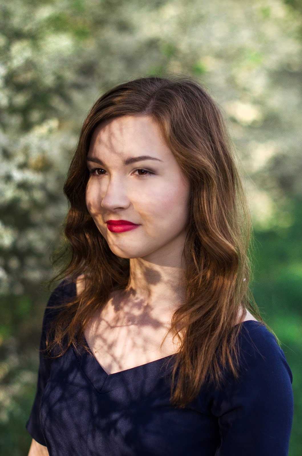 Portret młodej kobiety z długimi brązowymi włosami, czerwonymi ustami i ciemnoniebieską bluzką, w tle rozmyte kwitnące drzewo, twarz częściowo zacieniona przez gałęzie.