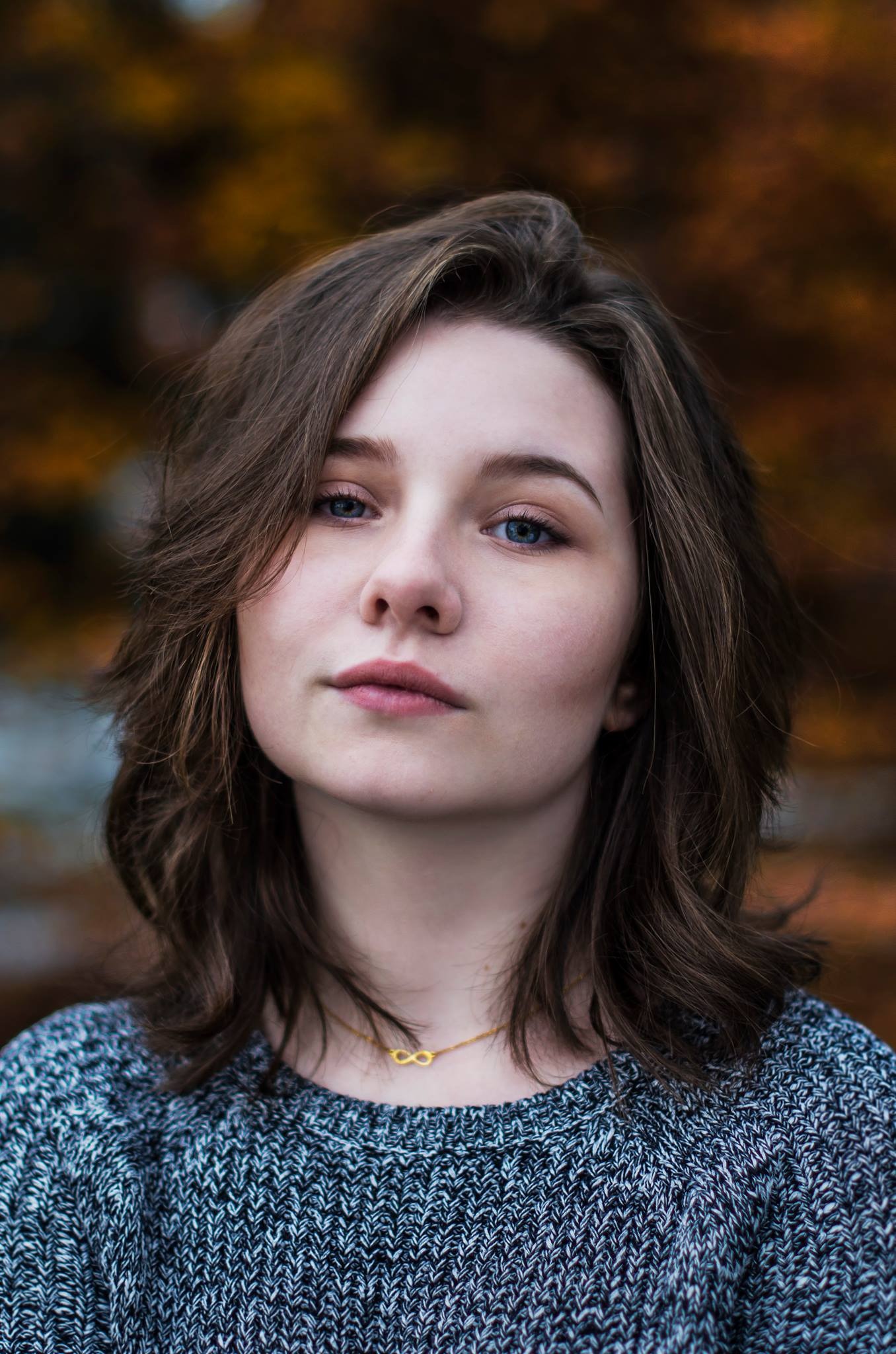 Portret młodej kobiety z niebieskimi oczami i złotym naszyjnikiem z symbolem nieskończoności na tle jesiennych liści.