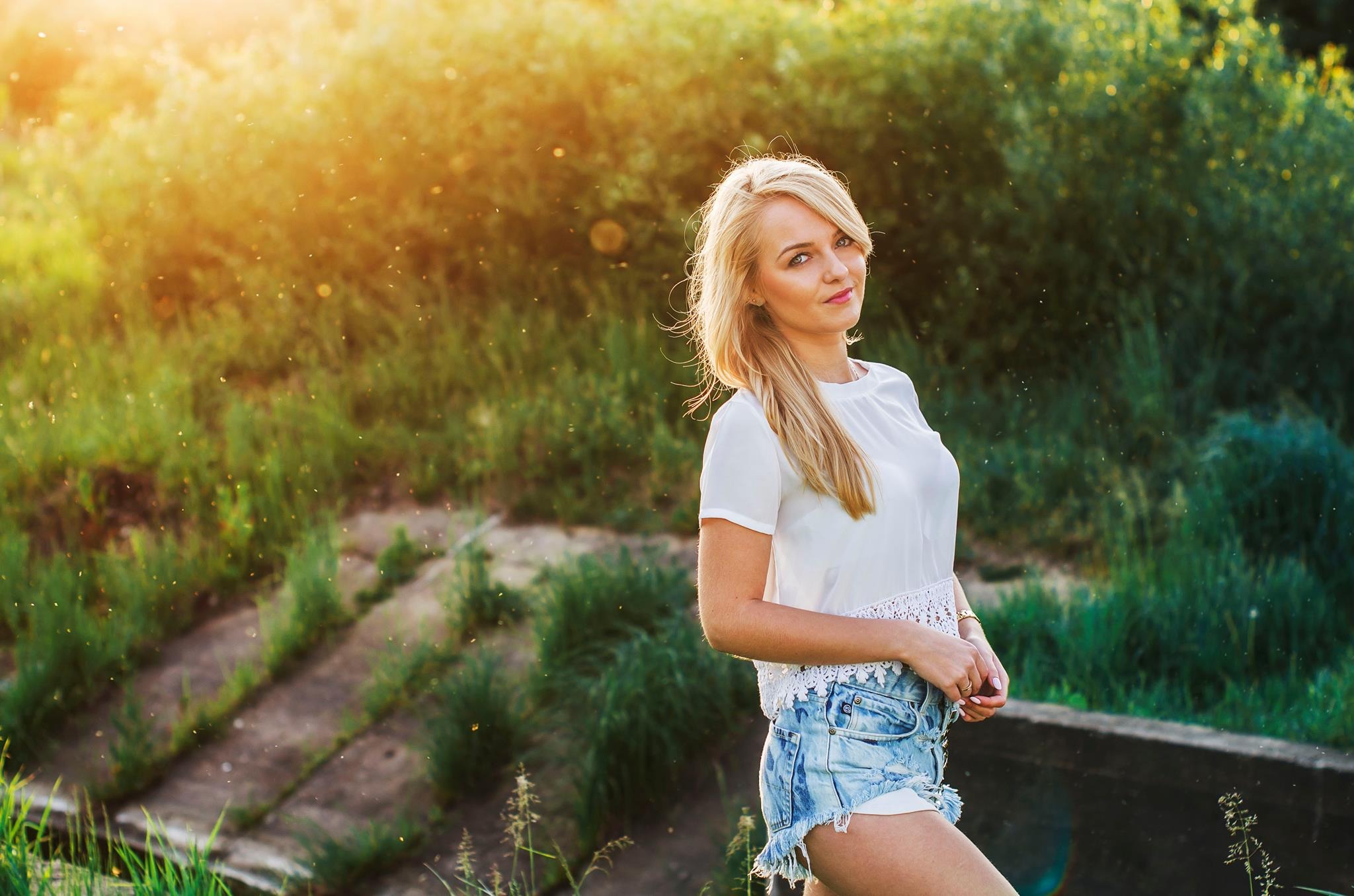 Portret uśmiechniętej blondynki w białej bluzce i jeansowych szortach na tle zielonej roślinności i zachodzącego słońca.