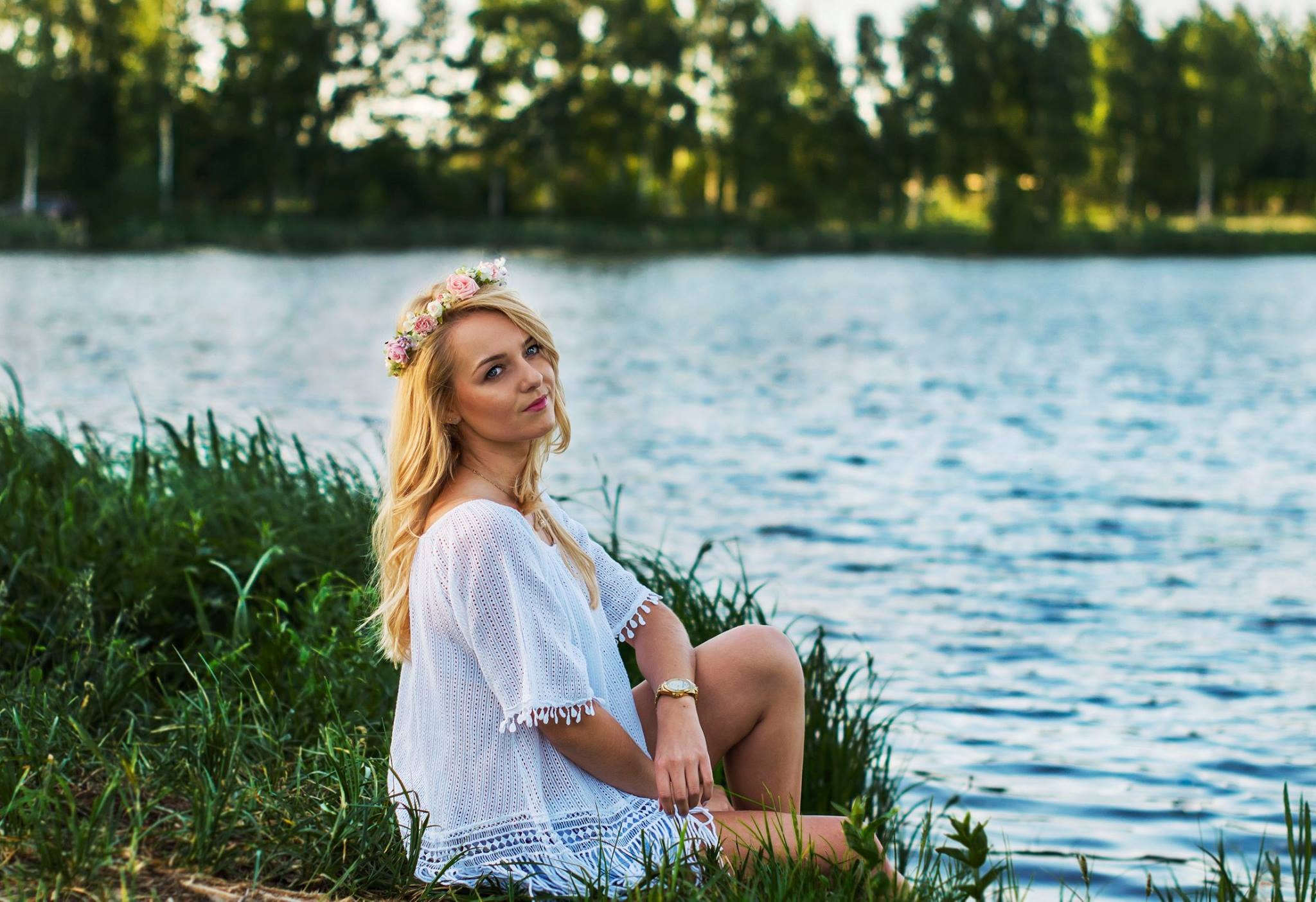 Portret kobiety z blond włosami i wiankiem z róż siedzącej na trawie nad brzegiem jeziora, ubrana w białą, ażurową bluzkę, z delikatnym uśmiechem i złotym zegarkiem na nadgarstku.