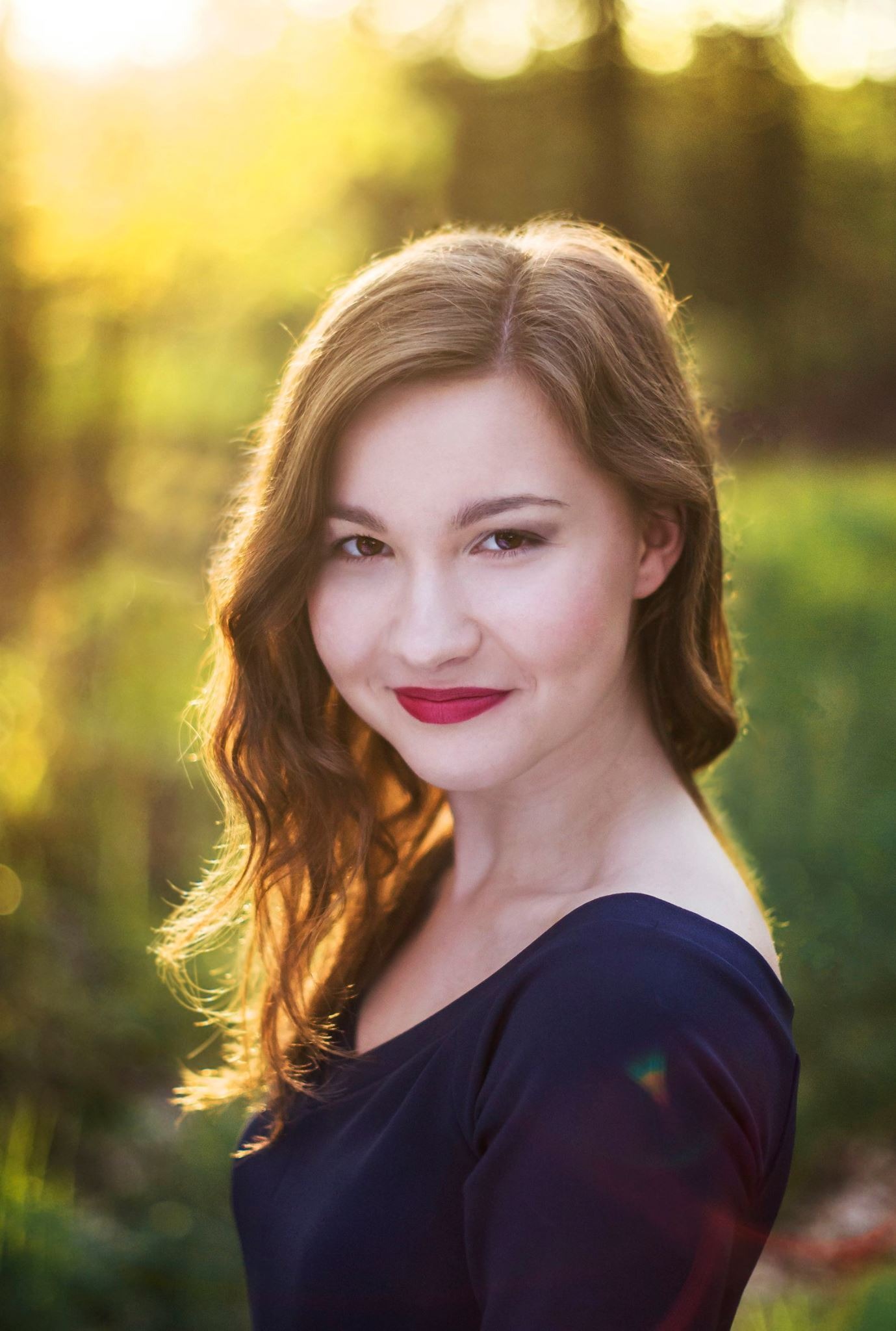 Portret uśmiechniętej młodej kobiety z rudymi, falowanymi włosami i ciemnoczerwoną szminką na ustach, w naturalnym świetle słonecznym.