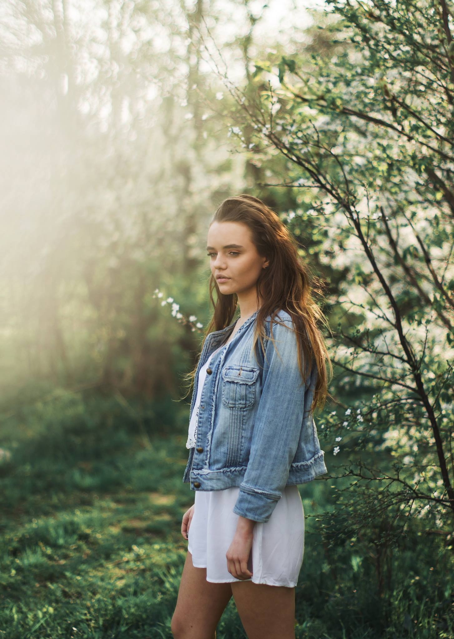 Portret młodej kobiety w dżinsowej kurtce i białej sukience, stojącej na tle kwitnących drzew w rozproszonym świetle.