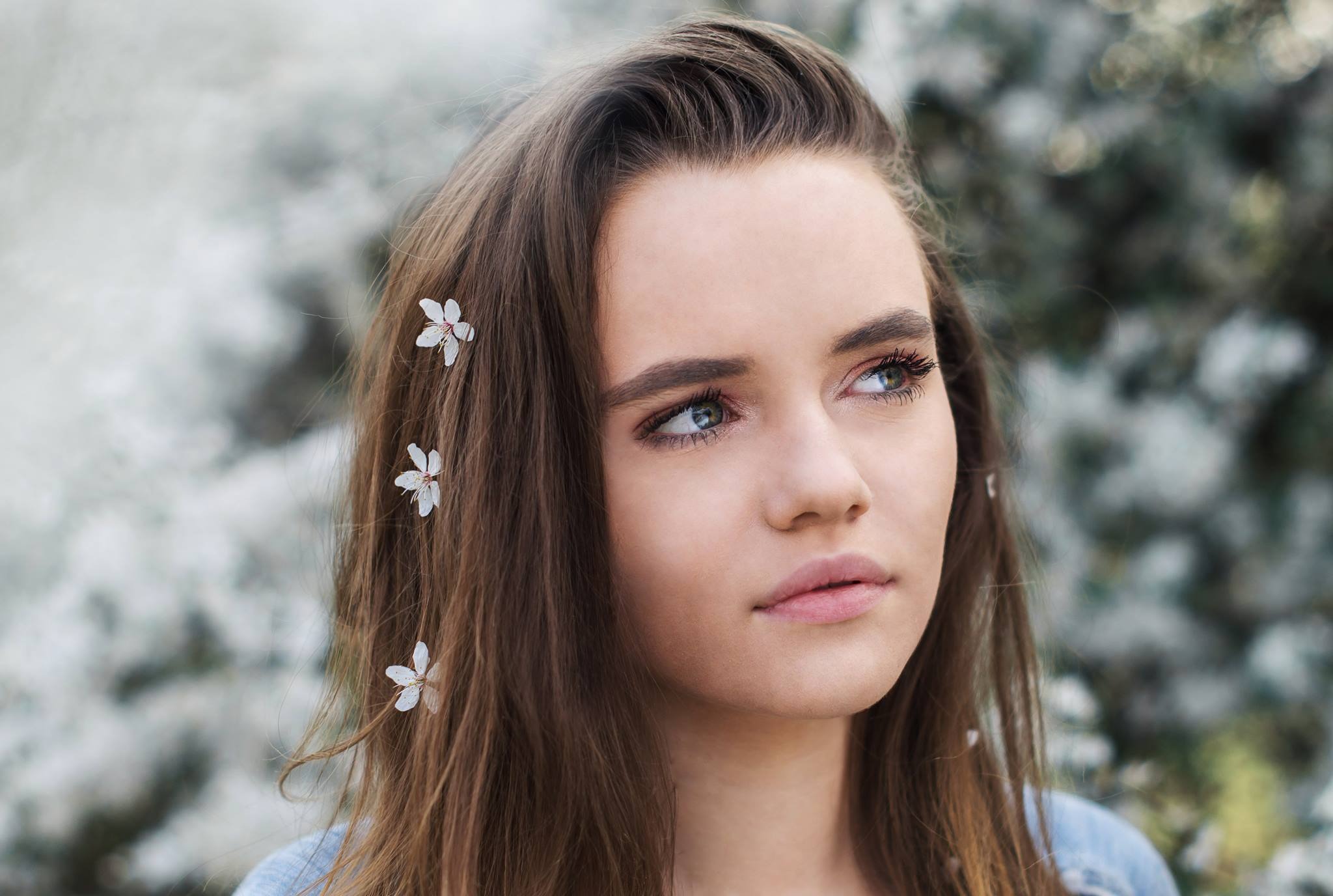 Portret młodej kobiety z kwiatami we włosach, patrzącej w górę na tle kwitnącego drzewa.