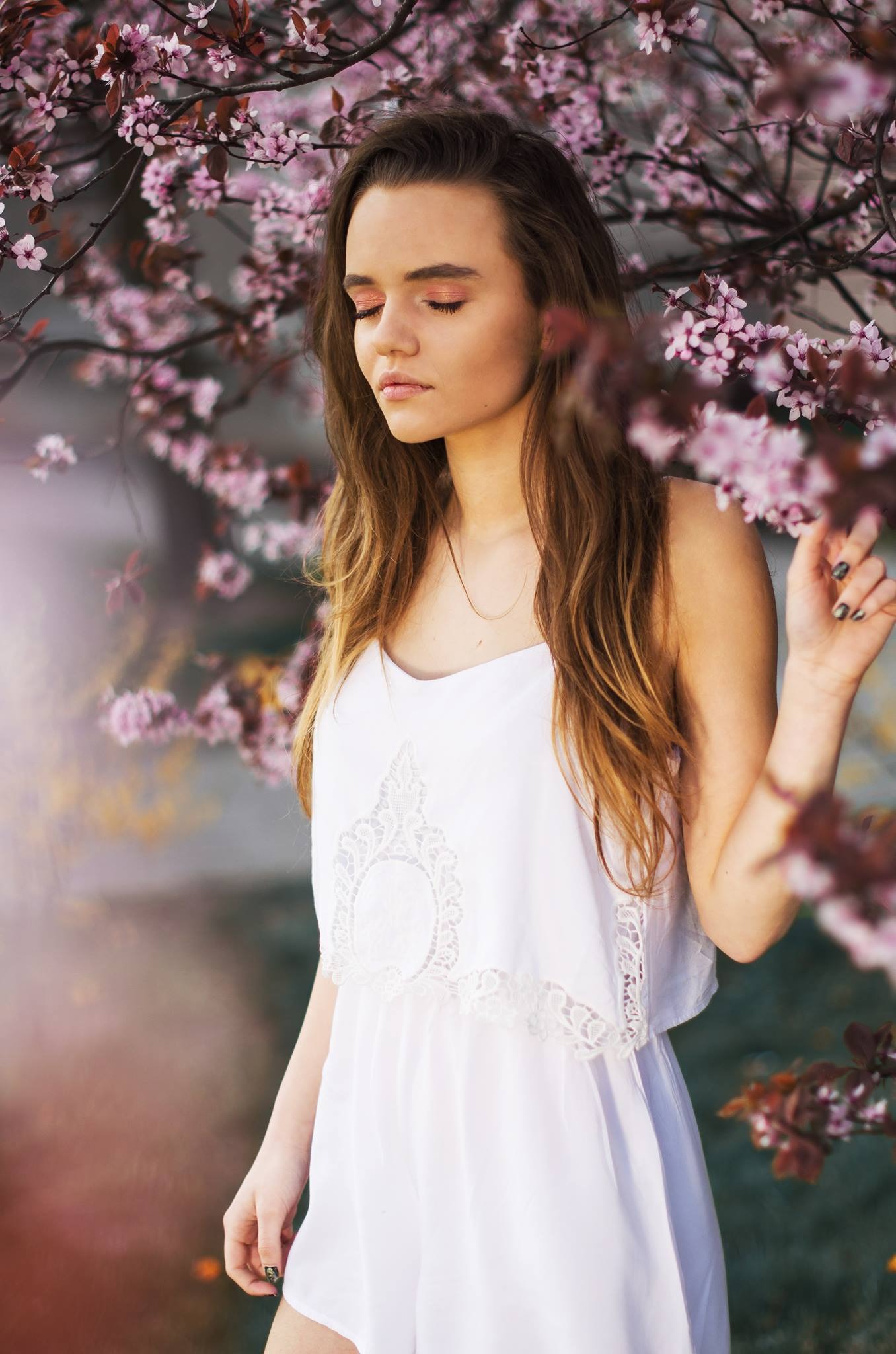 Portret młodej kobiety w białej sukience, z zamkniętymi oczami, na tle kwitnącej wiśni o różowych kwiatach, delikatny makijaż w odcieniach różu.