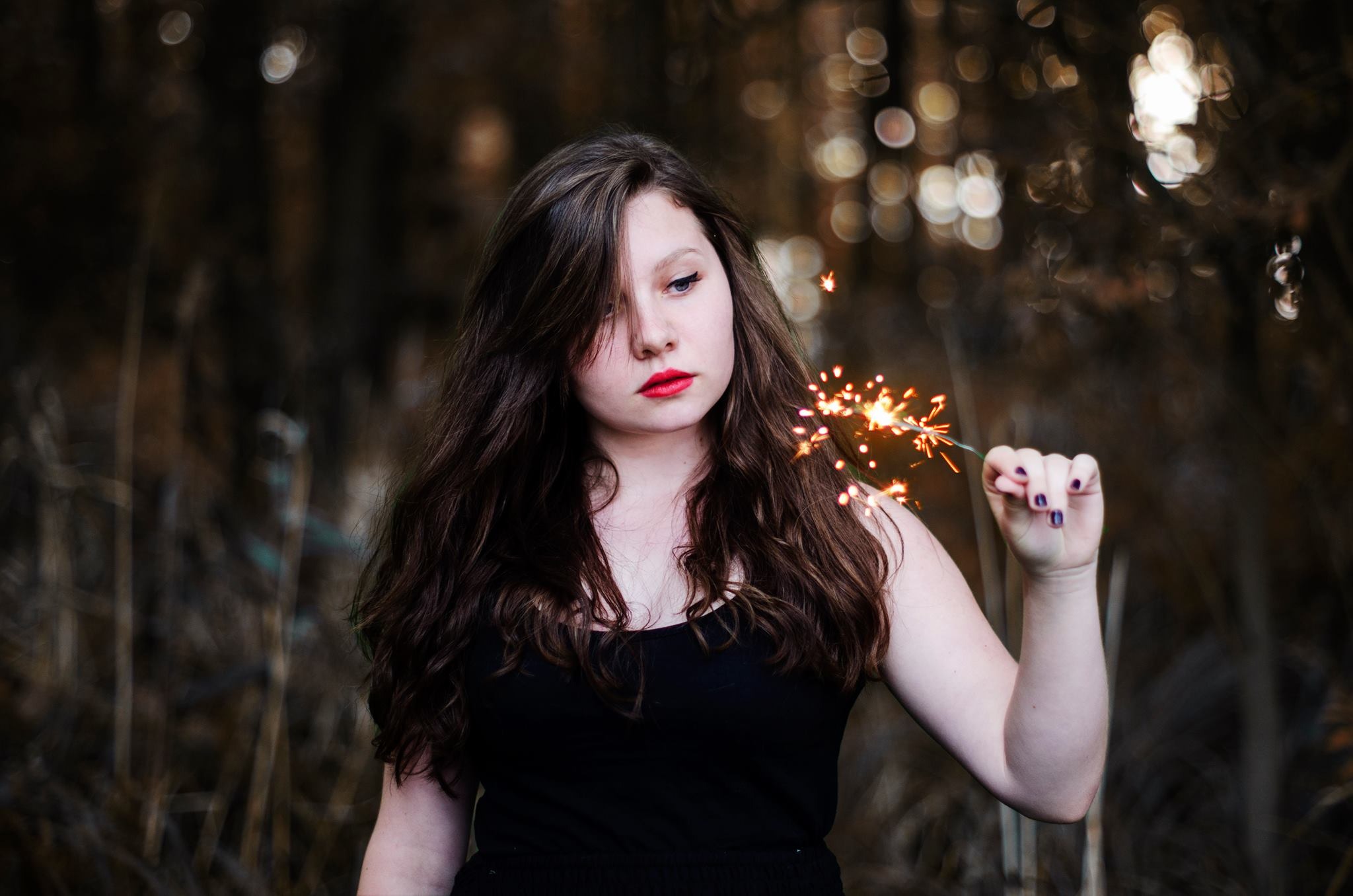Portret młodej kobiety z długimi brązowymi włosami, trzymającej zapaloną zimną iskrę na tle rozmytego, jesiennego krajobrazu. Czerwone usta i ciemny lakier na paznokciach dodają wyrazistości.
