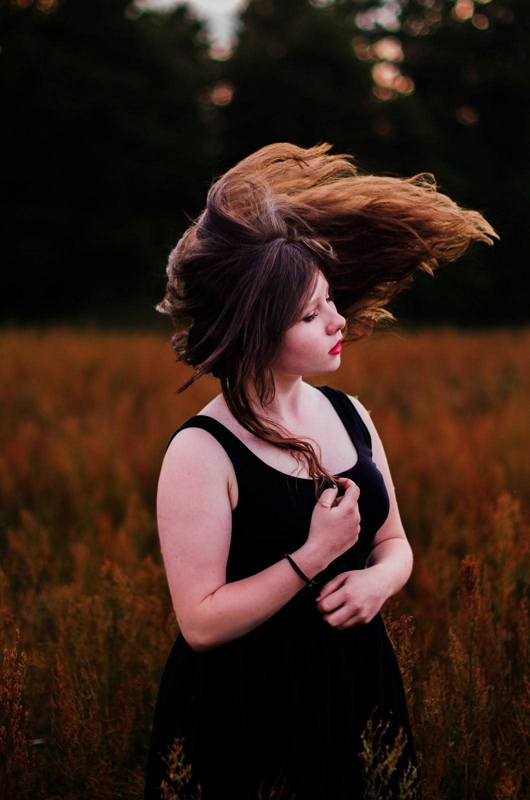 Portret kobiety w czarnej sukience, odwracającej głowę z rozwianymi włosami na tle pola traw w odcieniach brązu.