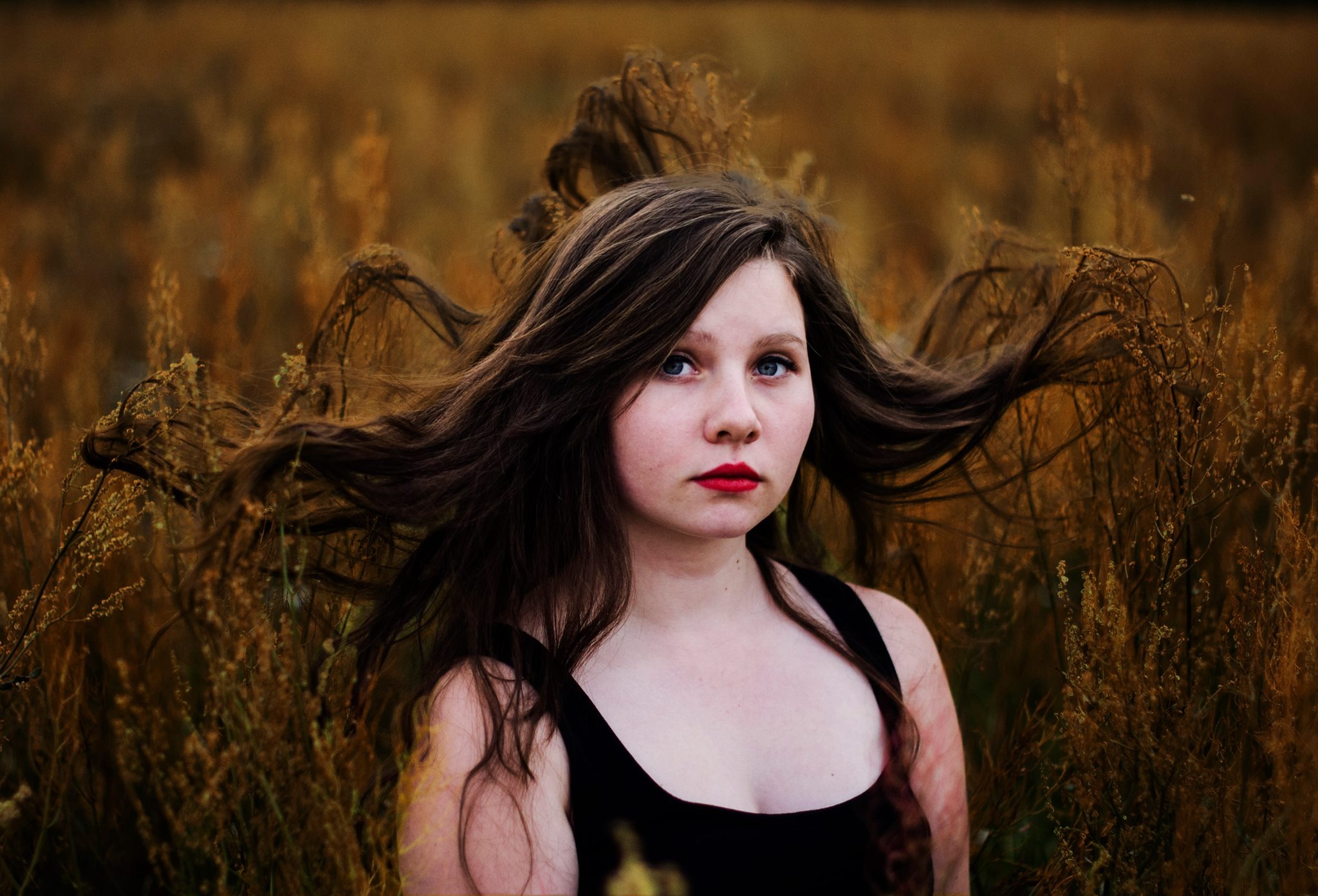 Portret młodej kobiety z rozwianymi brązowymi włosami i czerwonymi ustami na tle pola z wysoką, suchą trawą.
