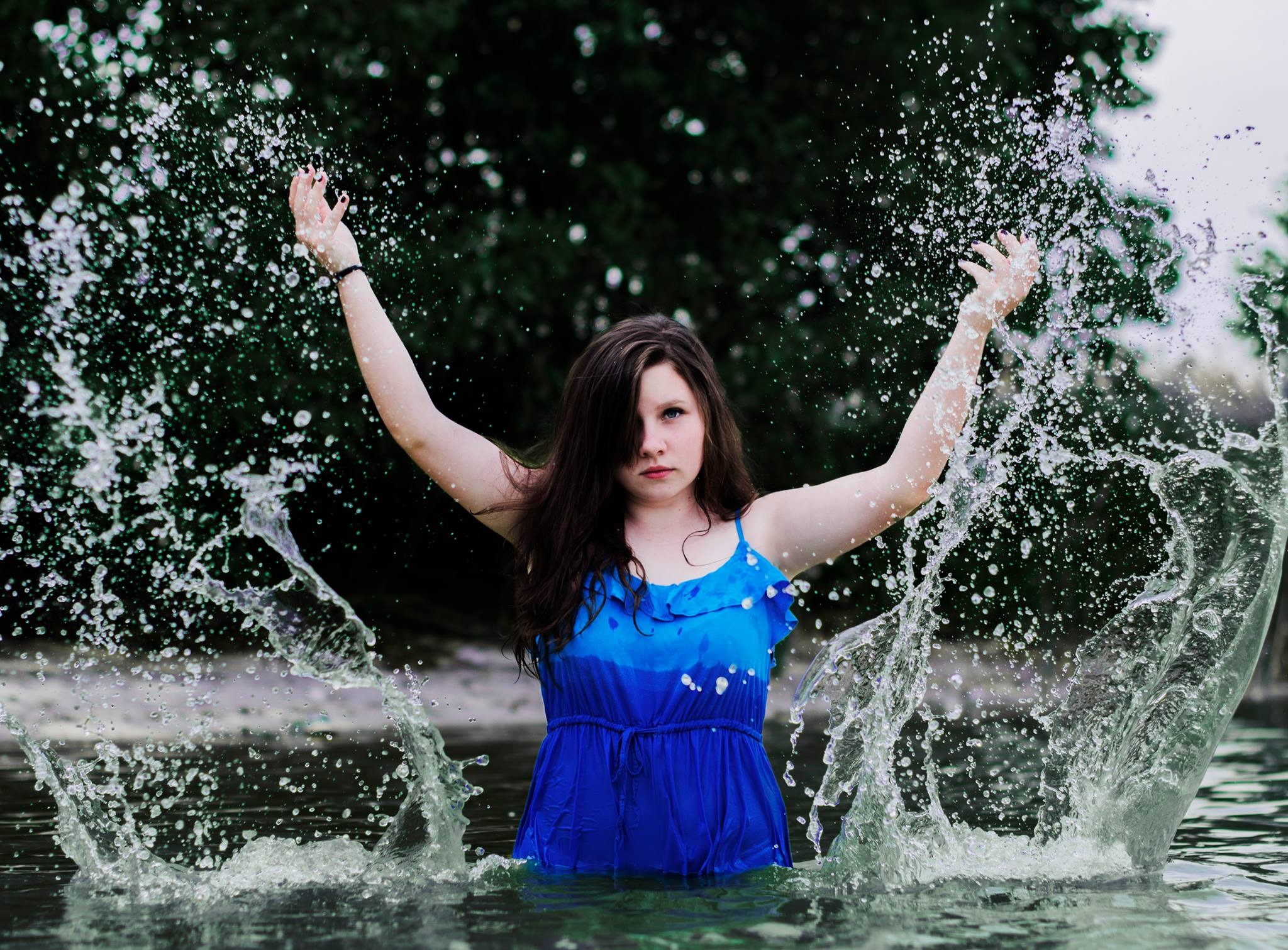 Młoda kobieta w niebieskiej sukience stoi w wodzie, unosząc ręce, otoczona rozpryskującą się wodą na tle zieleni.