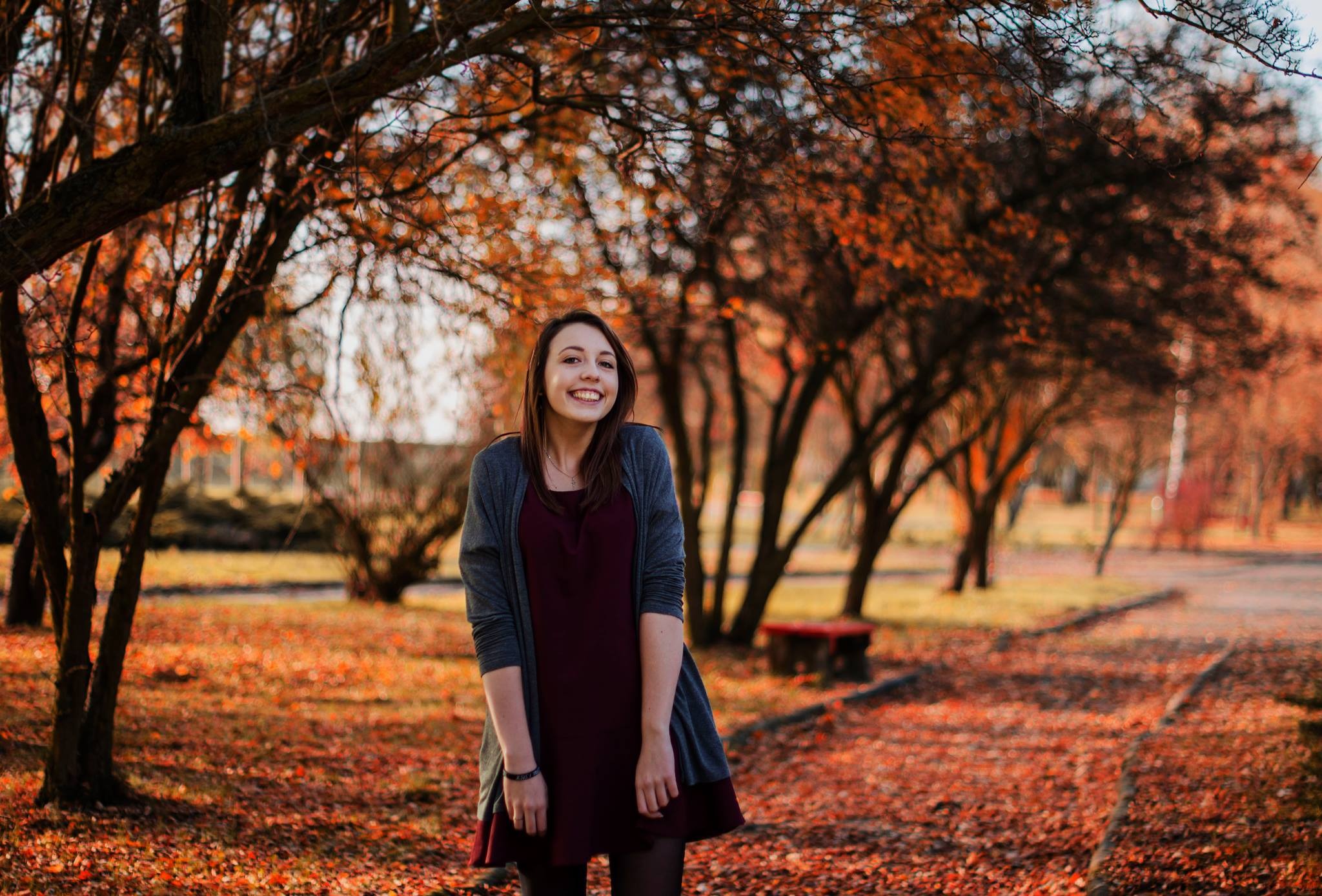 Portret uśmiechniętej młodej kobiety w bordowej sukience i szarym kardiganie na tle jesiennego parku z opadłymi liśćmi.