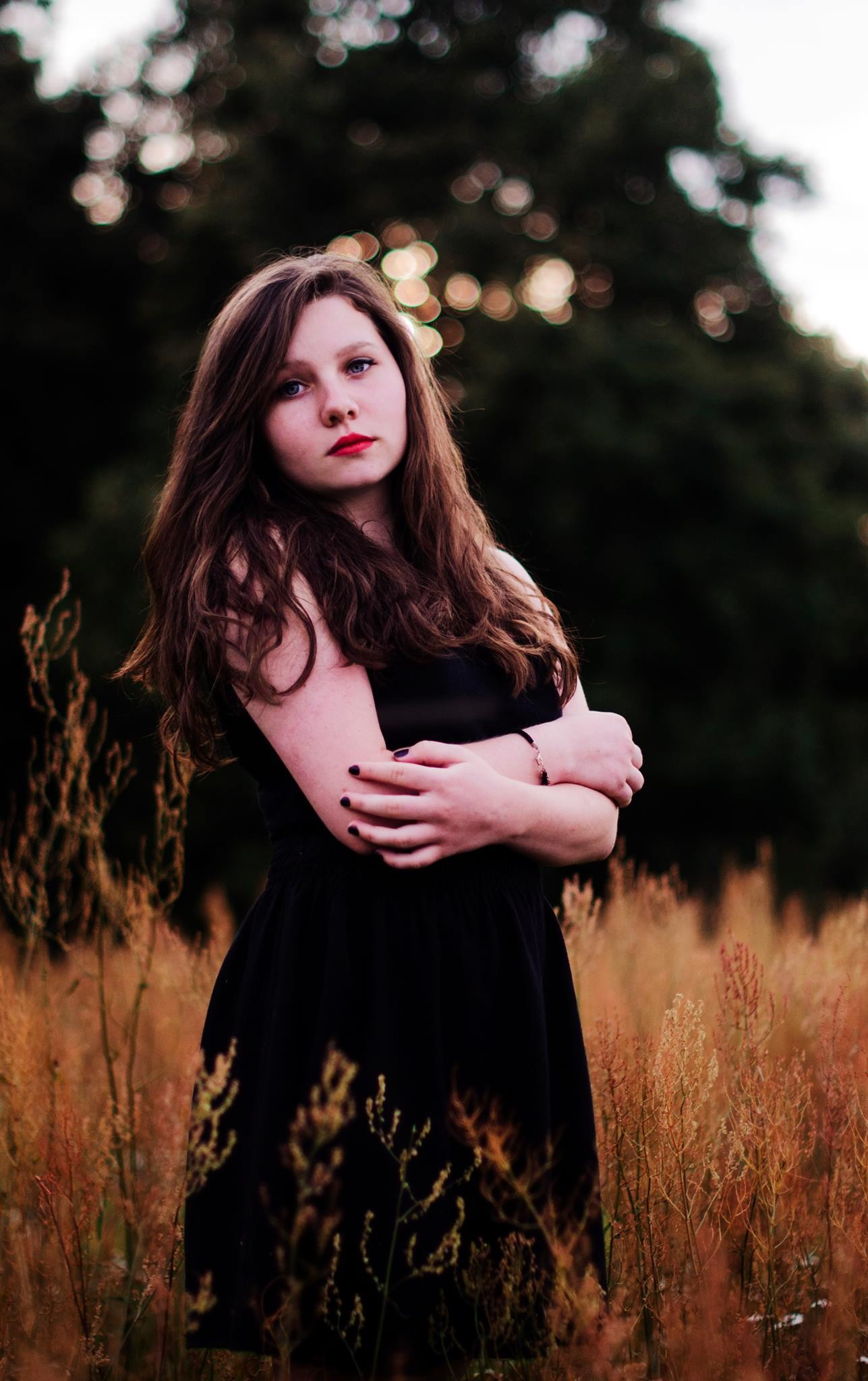 Portret młodej kobiety w czarnej sukience, stojącej na łące z wysoką trawą, skrzyżowane ręce, naturalny makijaż i rozpuszczone włosy, ciemne tło z bokeh.