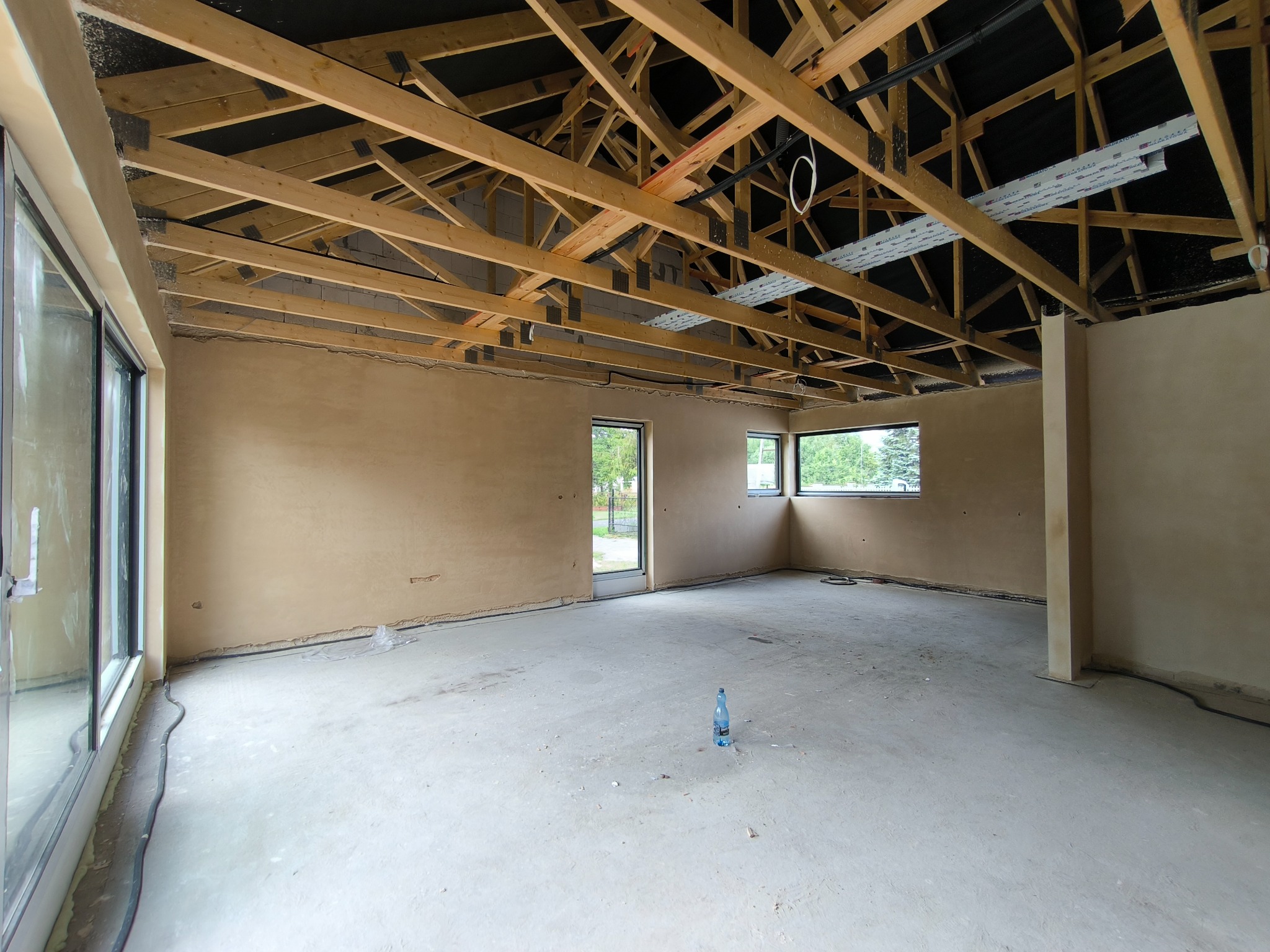 Surowe, jasne wnętrze w trakcie tynkowania maszynowego, z widoczną drewnianą konstrukcją dachu i butelką wody na posadzce.