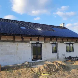 Niedokończony dom z białego pustaka z zainstalowanymi panelami słonecznymi na czarnym dachu, widoczne okna z czarnymi ramami i sterta piasku na pierwszym planie.