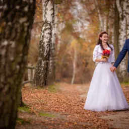 Para młoda, idąca ścieżką w lesie brzozowym, trzymająca się za ręce; panna młoda w białej sukni i z bukietem czerwonych kwiatów, pan młody w niebieskim garniturze w kratę i bordowej muszce.